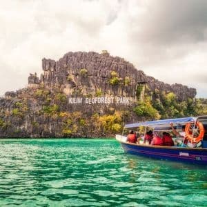 Eine WeRoad-Gruppenreise auf einem Boot segelt auf türkisfarbenem Wasser und nähert sich dem Schild des Kilim Geoforest Parks auf einer felsigen Insel.