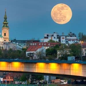 Una grande luna piena si staglia nel cielo crepuscolare sopra uno skyline cittadino, con un campanile e un ponte illuminato in primo piano.