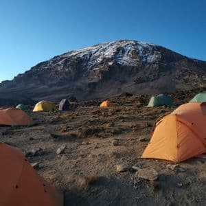 Tende colorate punteggiano un campeggio roccioso ai piedi di una montagna innevata sotto un cielo azzurro e limpido.