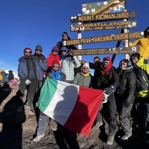 Un gruppo WeRoad in viaggio posa con la bandiera italiana sulla vetta Uhuru Peak del Monte Kilimanjaro, sopra un mare di nuvole.