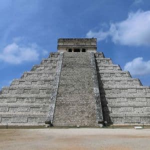 Una grande piramide a gradoni in pietra, con una scalinata centrale, si staglia al centro dell'immagine sotto un cielo blu con nuvole bianche.