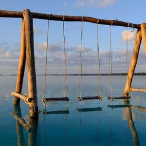 Un'altalena di legno, con diversi seggiolini vuoti, si erge su un'acqua calma e limpida, il suo riflesso visibile in superficie.