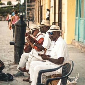 Three musicians in white outfits and hats sit on a cobblestone street playing guitars and other instruments.