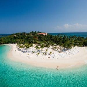 An aerial view of a tropical island with a white sand beach, palm trees, and lounge chairs, surrounded by clear turquoise water.