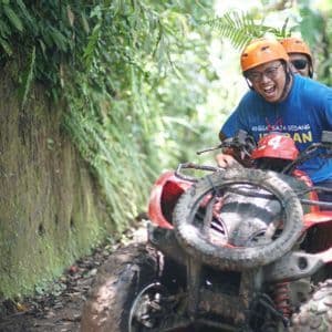 Two people on a WeRoad group trip laughing while riding a muddy red quad bike on a trail through a lush jungle.