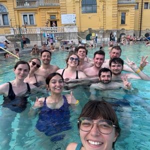 Eine WeRoad-Gruppe macht lächelnd ein Selfie in einem Freibad vor einem großen, gelben historischen Gebäude.