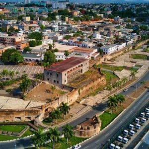 An aerial view of a historic coastal city showing ancient stone fortifications, a large plaza, and a modern road alongside the water.