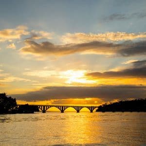 A silhouetted arched bridge crosses a body of water as the sun sets, casting a golden glow on the clouds and water.