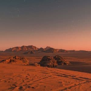A vast desert landscape with red sand and rocky mountains under a starry sky at dusk.