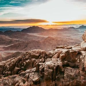 The sun rises over a vast, hazy mountain range, viewed from a rocky summit under a turquoise and orange sky.