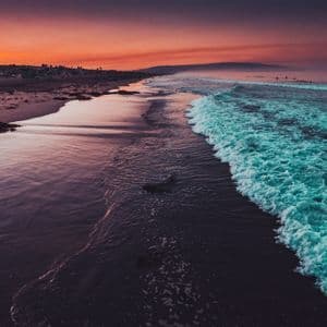 Turquoise waves roll onto a dark sand beach, reflecting the vibrant orange and purple colors of the sunset sky.