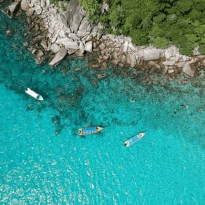 Vista aerea di tre barche che galleggiano in acqua turchese mentre persone nuotano e fanno snorkeling lungo una costa rocciosa e boscosa.