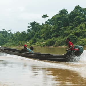 Tre persone su una lunga canoa a motore navigano lungo un fiume, con una persona che indica la fitta giungla sulla riva.
