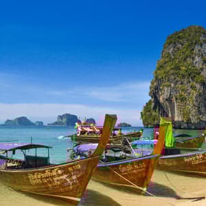 Barche a coda lunga thailandesi ormeggiate su una spiaggia sabbiosa in acque turchesi, con faraglioni calcarei sullo sfondo sotto un cielo azzurro.