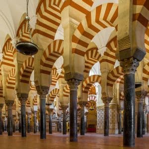 Fila di colonne scure sorreggono doppi archi a strisce rosse e bianche in una grande sala con un alto soffitto a volta e lampade a sospensione.