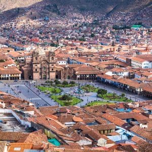 An aerial view of a bustling city square surrounded by historic buildings with terracotta roofs, with mountains visible in the background.