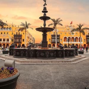 An ornate bronze fountain stands in a bustling city plaza surrounded by yellow colonial buildings and palm trees at sunset.