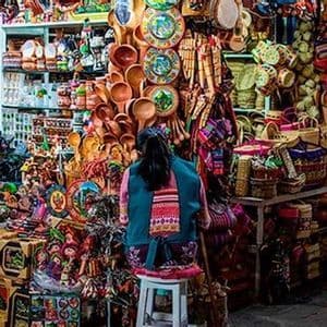 A woman with her back to the camera sits on a stool in a busy market stall filled with colorful handmade souvenirs and crafts.
