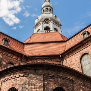 Un'inquadratura dal basso di una chiesa storica in mattoni, con un tetto di tegole rosse e una torre dell'orologio, su un cielo azzurro con nuvole.