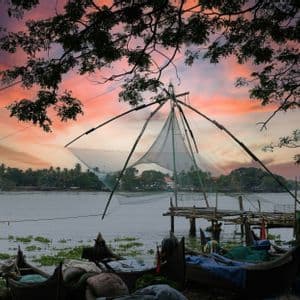 Una grande rete da pesca cinese si erge sulla riva di un lago, accanto a diverse barche ormeggiate, sotto un cielo colorato al tramonto e incorniciata da rami d'albero.
