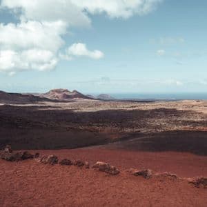 Un vasto paesaggio vulcanico con terreno rosso in primo piano si estende verso montagne distanti e l'oceano sotto un cielo nuvoloso.