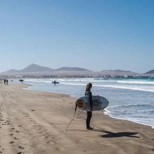 Un surfista in muta tiene una tavola da surf su una spiaggia sabbiosa, guardando l'oceano con montagne distanti sotto un cielo sereno.
