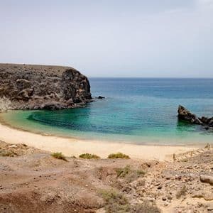 Panoramica da una scogliera rocciosa su una spiaggia sabbiosa appartata, incastonata in una caletta con acque turchesi e cristalline, sotto un cielo leggermente velato.