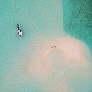 Vista aerea di una piccola barca bianca che galleggia in acqua turchese cristallina, vicino a un banco di sabbia con due persone.