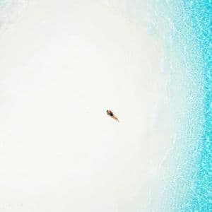 Vista aerea di una persona in costume da bagno sdraiata su una spiaggia di sabbia bianca accanto ad acqua turchese cristallina.