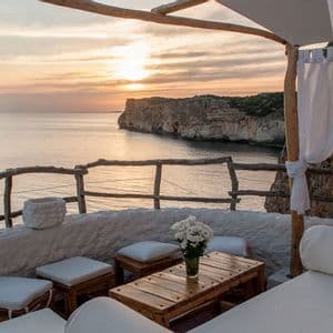 A cliffside terrace with wooden furniture and white flowers overlooks the calm sea and a rocky coastline during sunset.