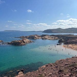 Una vista dall'alto di una caletta rocciosa con acqua turchese, dove le persone nuotano e si rilassano su una piccola spiaggia sabbiosa.