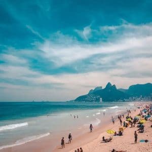 A crowded sandy beach with people sunbathing under umbrellas and swimming in the ocean, with a city and mountains in the background.