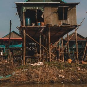 Un villaggio di case rustiche su palafitte sulla riva di un fiume, con una grande casa di lamiera ondulata in primo piano e bambini sotto.