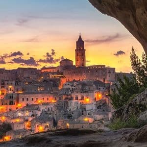 Un'antica città collinare con un campanile imponente, illuminata da luci calde al tramonto e vista dall'interno di una grotta.