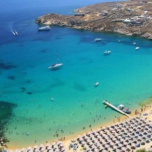 Vista aerea di una spiaggia sabbiosa affollata con ombrelloni, accanto a una baia turchese con bagnanti e barche.