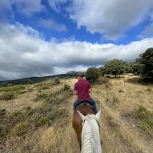 Una persona in camicia rossa cavalca un cavallo attraverso un paesaggio collinare ed erboso sotto un cielo azzurro e nuvoloso, vista dalla groppa di un altro cavallo.