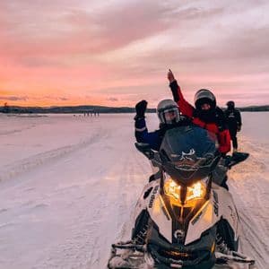 Deux personnes d'un voyage de groupe WeRoad saluent depuis une motoneige dans un vaste paysage enneigé sous un ciel de coucher de soleil rose.