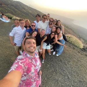 Un voyage de groupe WeRoad souriant pour un selfie pris par un homme en chemise rose sur une pente volcanique au coucher du soleil.