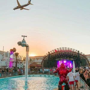 Un pubblico a un festival in piscina osserva un palco al tramonto, con una scultura di rubinetto galleggiante e un aereo a bassa quota.