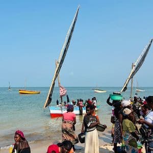 Eine Gruppe Einheimischer versammelte sich an einem Sandstrand neben traditionellen Segelbooten auf dem Wasser unter einem klaren blauen Himmel.
