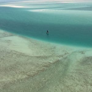 Vista aerea di una singola barca a vela su vaste acque turchesi e poco profonde con banchi di sabbia in lontananza.