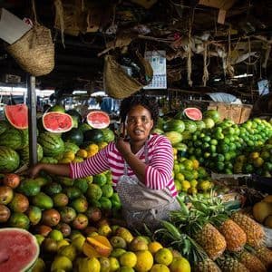 Una fruttivendola sorride dietro la sua bancarella del mercato, che trabocca di angurie, manghi e ananas.