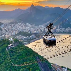 A cable car is suspended high above a lush green mountain, overlooking a cityscape and a bay filled with boats at sunset.