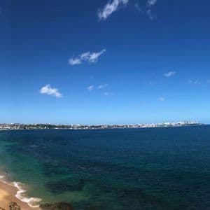 Una vista panoramica dall'alto di una costa cittadina con una lunga spiaggia di sabbia, acque turchesi e un cielo azzurro limpido con qualche nuvola.