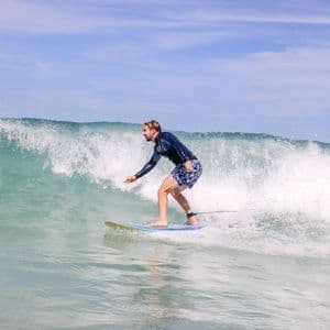 Un uomo con una rash guard e pantaloncini si equilibra su una tavola da surf mentre cavalca un'onda dell'oceano sotto un cielo parzialmente nuvoloso.
