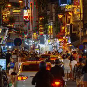 Una grande folla di persone cammina lungo una stretta via di città di notte, piena di auto, moto e luminose insegne al neon colorate.