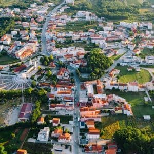 Una veduta aerea di un paesino con edifici dai tetti rossi, una piazza circolare e alberi verdi immersi nella calda luce del sole.