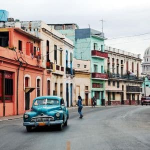 Une voiture vintage bleue roulant dans une rue bordée de bâtiments colorés, avec un grand bâtiment à dôme en arrière-plan.