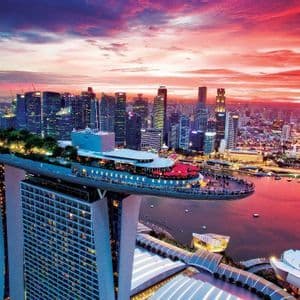 Vista aerea di un hotel moderno con terrazza panoramica affacciata su un denso skyline cittadino e una baia, durante un drammatico tramonto rosso.