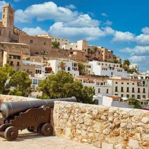 Un vieux canon repose sur un mur de pierre, dominant une ville historique à flanc de colline avec un clocher, sous un ciel bleu et des nuages blancs.